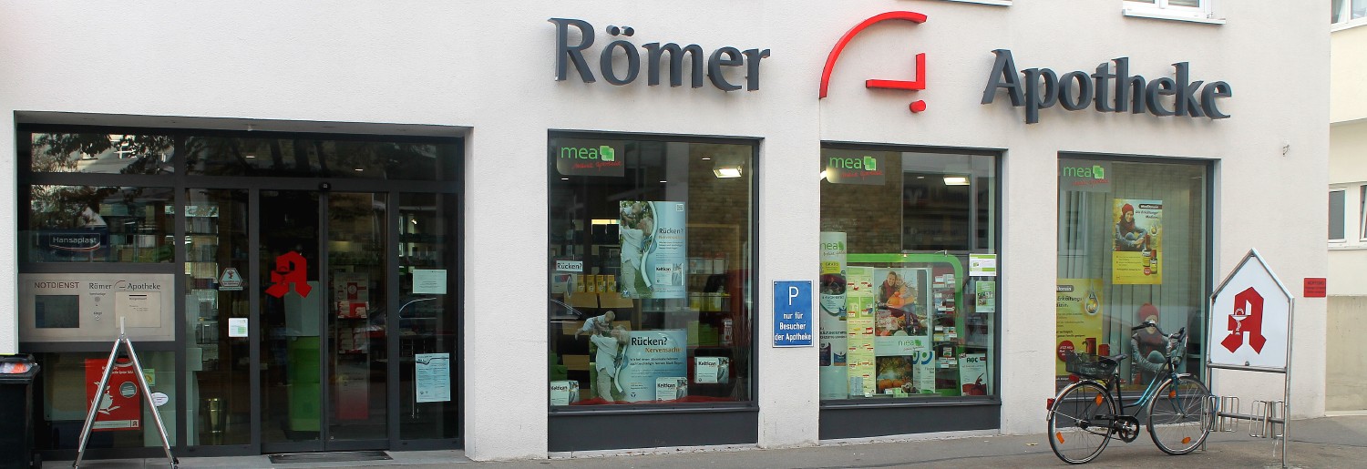 Römer Apotheke Ulm – Außenansicht mit Schaufenstern und Notdiensthinweis Außenansicht der Römer Apotheke in Ulm mit großer Glasfront, Apothekenschaufenstern und rotem Apotheken-A. Ein Fahrrad steht am Kundenständer, daneben befindet sich ein Hinweisschild zum Notdienst.